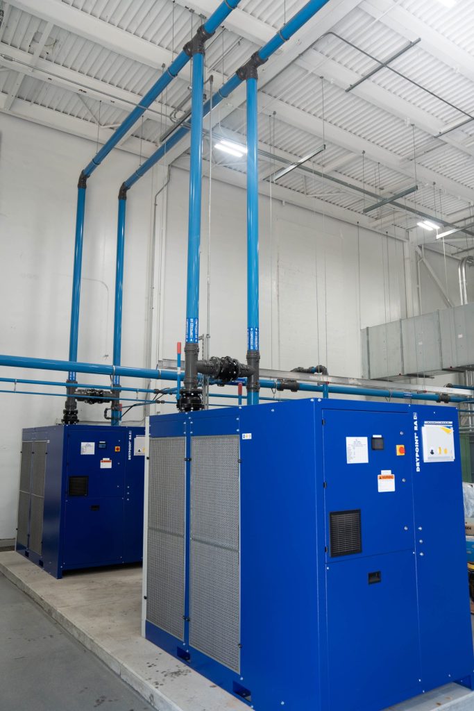 Two large blue industrial machines connected to overhead blue pipes in a clean, well-lit facility.
