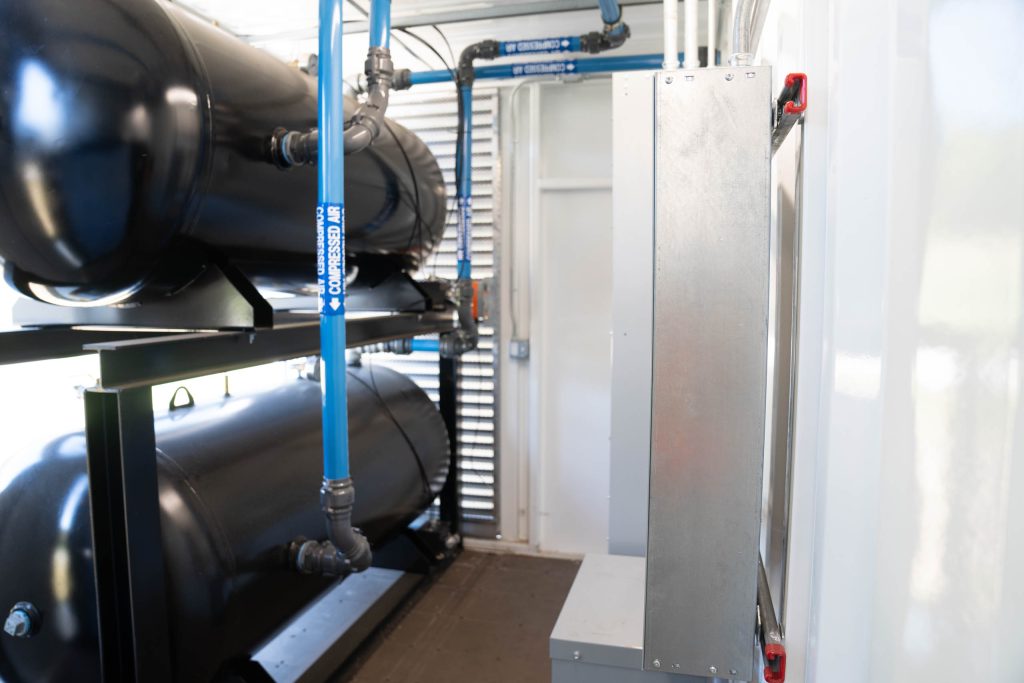 Large black cylindrical air tanks with blue labeled piping and metal control units inside an industrial facility.