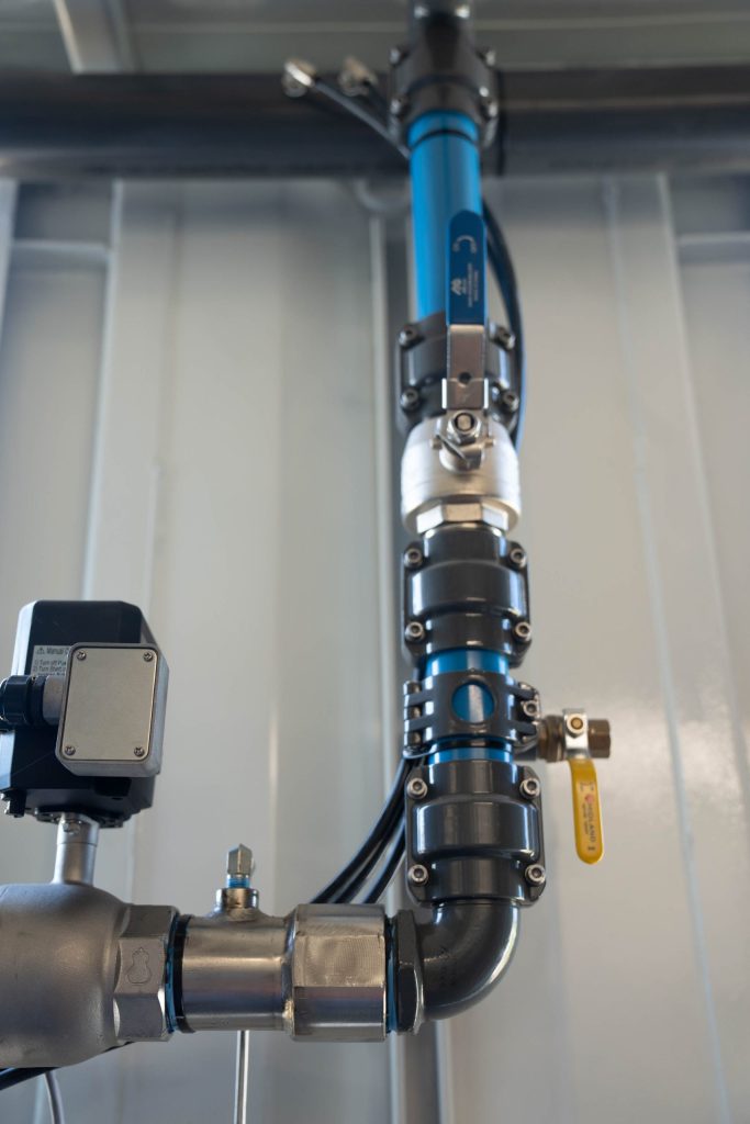Blue industrial pipeline with valve and sensor attached, mounted vertically against a corrugated metal wall.